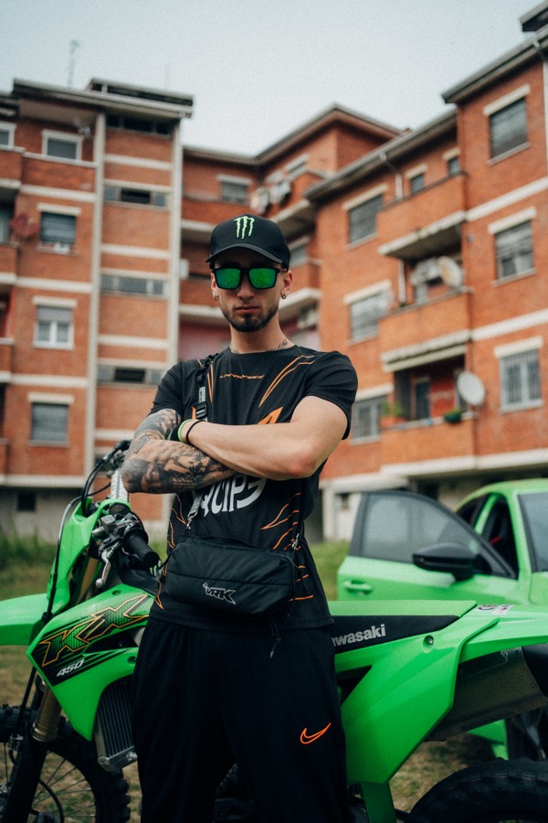 Azza, arms crossed, in front of the KX450 and red-brick housing blocks